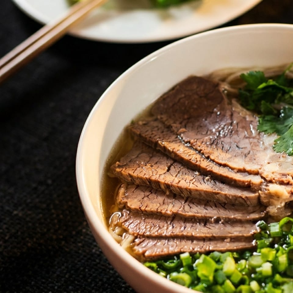 Close-up of a bowl of pho with sliced beef, noodles, cilantro, red onion, and green onions.