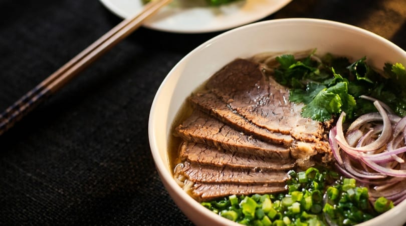 Close-up of a bowl of pho with sliced beef, noodles, cilantro, red onion, and green onions.
