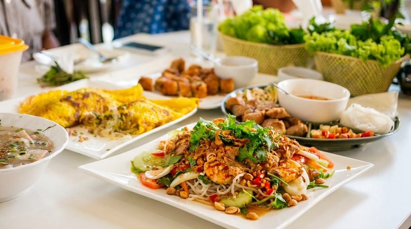 A table laden with various Vietnamese dishes, including a noodle salad, savory crepes, and spring rolls.